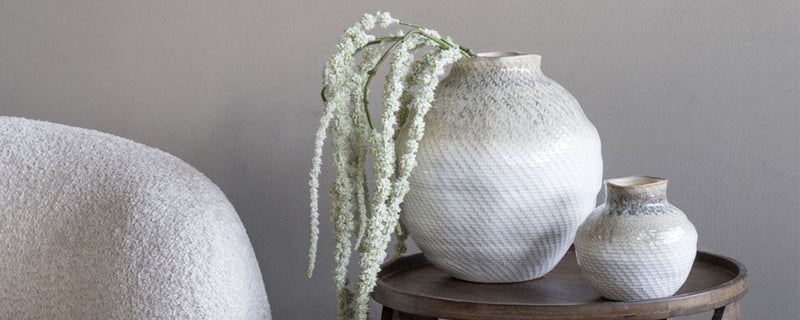 Two white flower vases sit on a table with dried flower stems