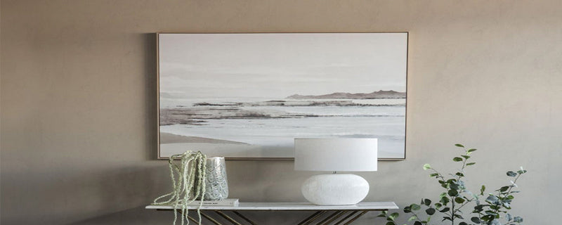 A large framed wall art landscape piece sits above a console table with a lamp and vase