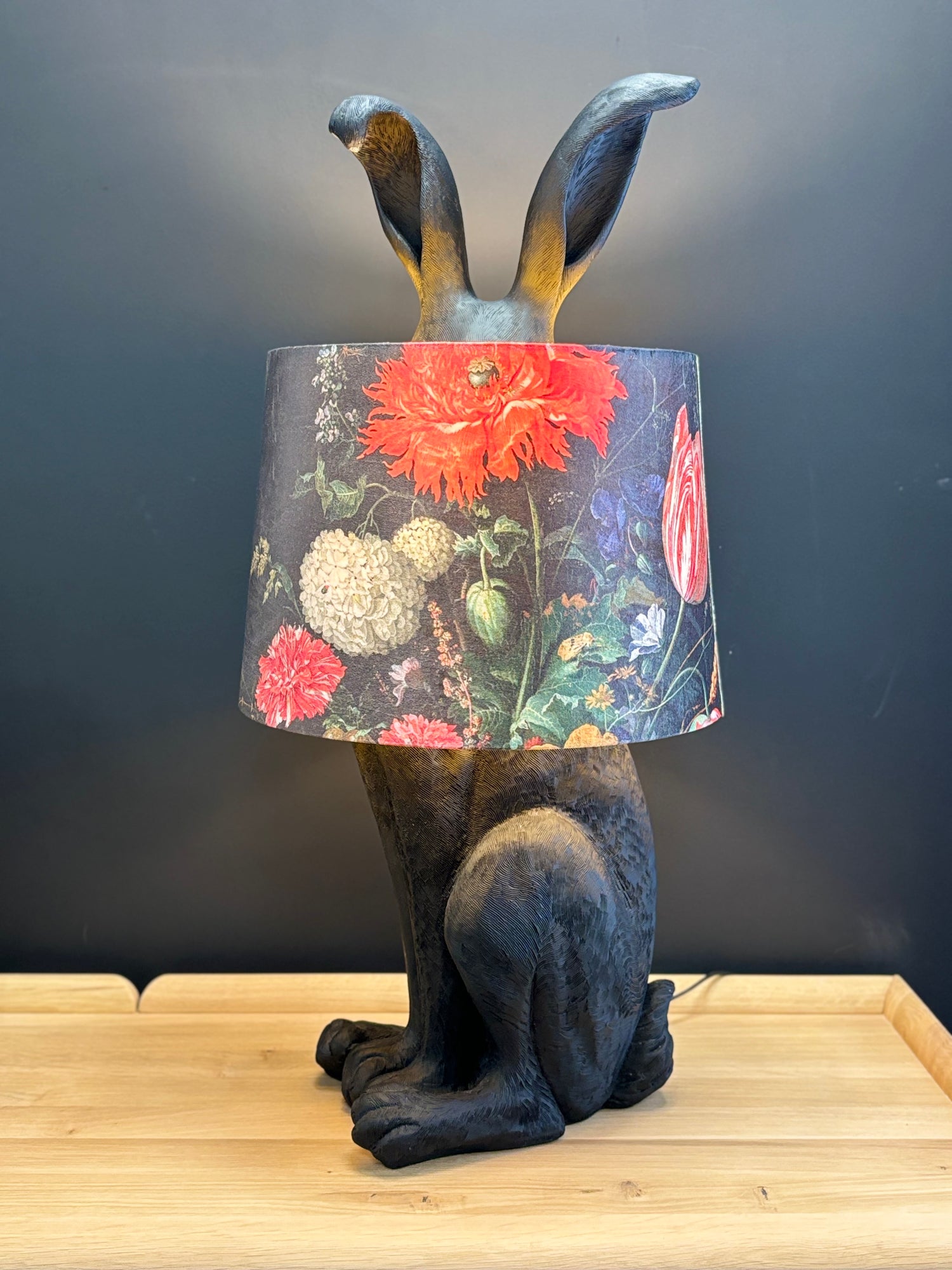 A sculptural rabbit or hare table lamp seated with a dark textured body and a cylindrical lampshade printed with a vintage-style floral pattern featuring red, pink, cream, and green flowers on a dark background.
