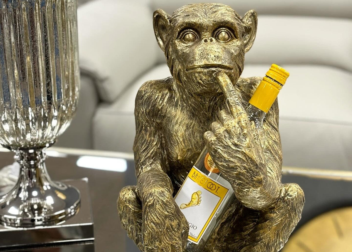 A metallic gold monkey bottle holder figurine sitting on a reflective surface, holding a small bottle with a yellow cap. 