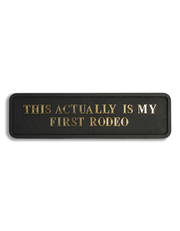 Black sign with gold text 'THIS ACTUALLY IS MY FIRST RODEO' 