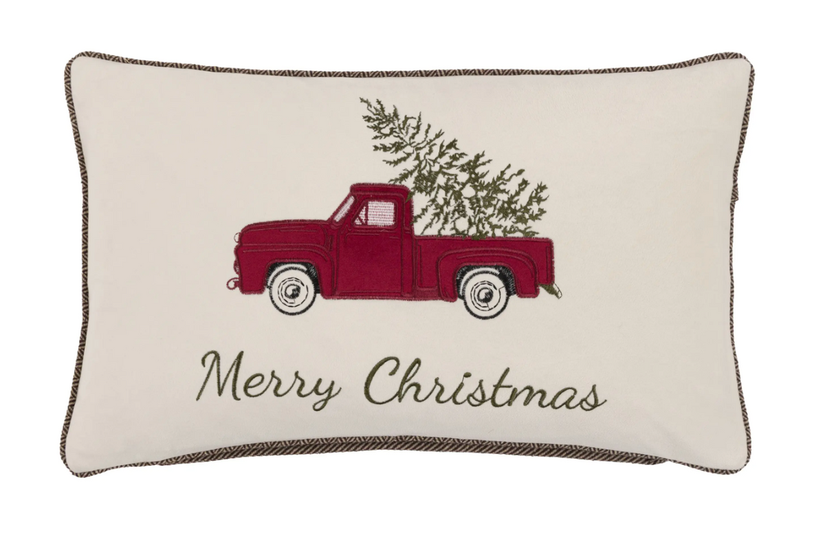 An off-white rectangular decorative throw pillow with a dark brown braided border, featuring an embroidered red vintage pickup truck carrying a green Christmas tree and the words 'Merry Christmas' in dark brown script.
