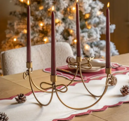 A four-arm brass candelabra holding four lit taper candles, styled on a table runner with pinecones and a blurred Christmas tree in the background.