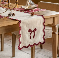 A cream table runner with scalloped burgundy edges and a bow motif, styled on a wooden dining table with chairs and christmas decor.